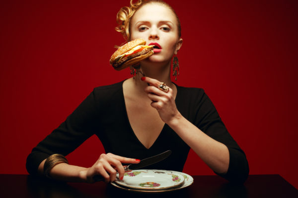 Fashion & Gluttony Concept. Portrait of luxurious red-haired model in black cocktail dress trying to eat burger over red background. Perfect hair, skin, make-up and manicure. Golden accessories. Studio shot